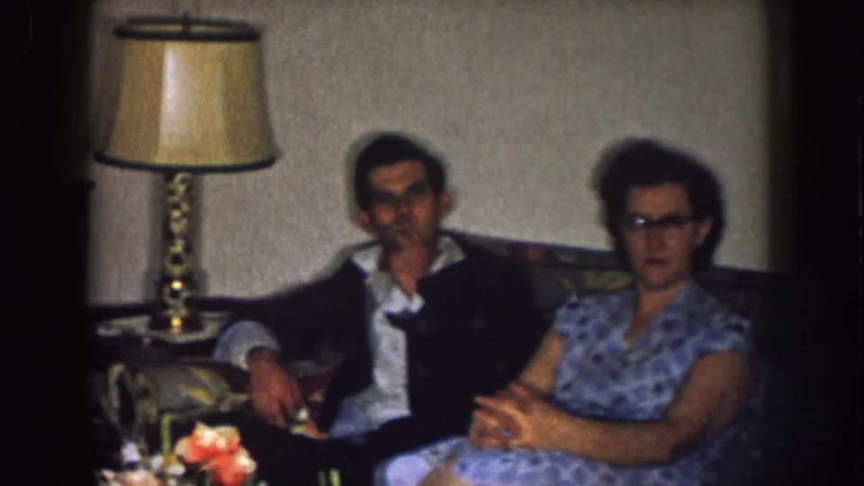 In the 1957 Nebraska living room footage, a man in a dark jacket and a woman in a blue patterned dress sit on a couch near a lamp.