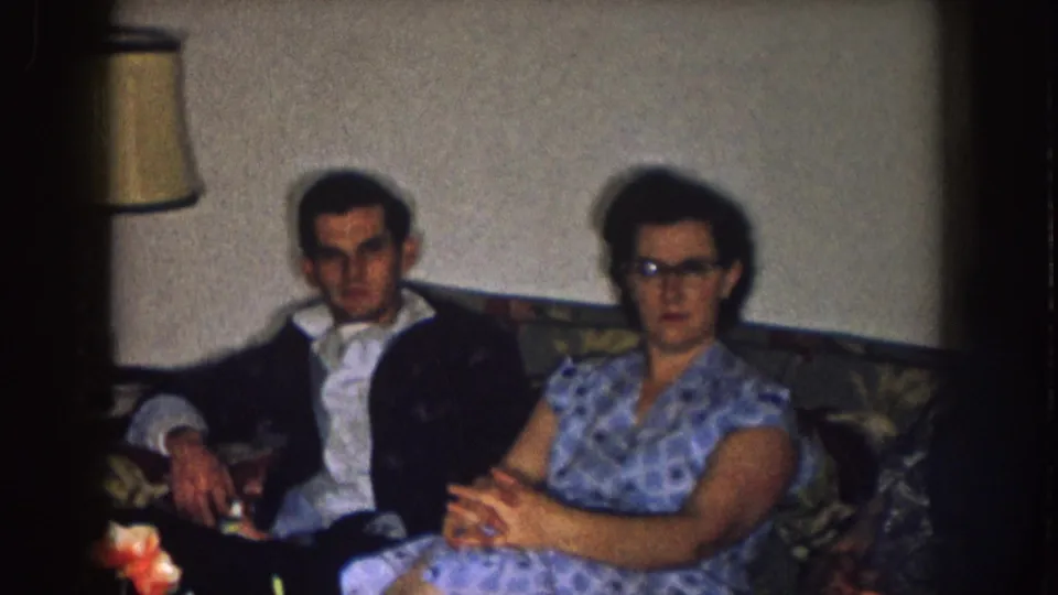 A man in a dark jacket and a woman in a blue patterned dress sit on a couch in a private Nebraska living room in 1957.