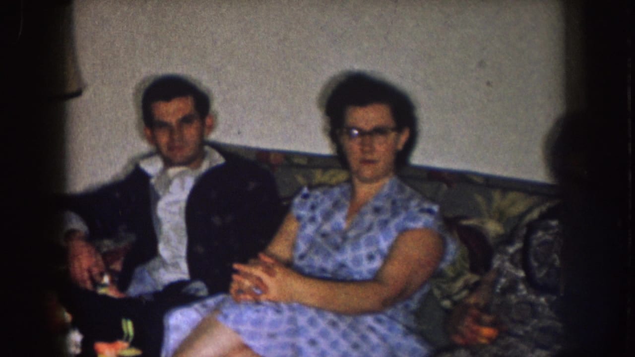 In a 1957 Nebraska home-movie frame, a man in a dark jacket and a woman in a blue patterned dress sit on a floral sofa.
