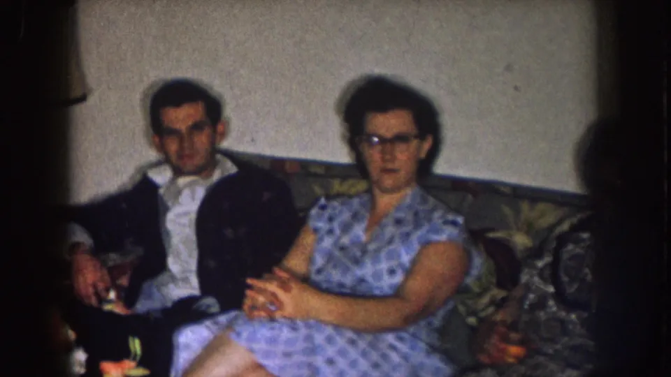 In a 1957 Nebraska home-movie frame, a man in a dark jacket and a woman in a blue patterned dress sit on a floral sofa.