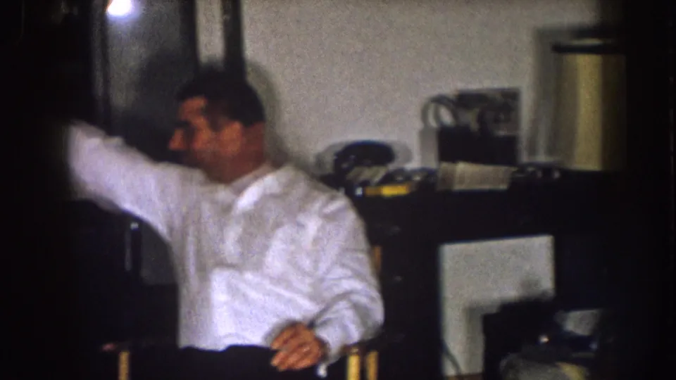 A man in a white shirt sits in the same private living room, with a table lamp and furniture visible behind him.