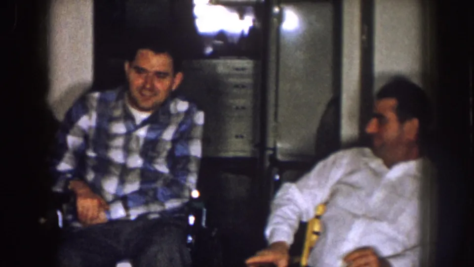 Two adult men sit in a private Nebraska living room, one in a plaid shirt and one in a white shirt, under warm indoor light.