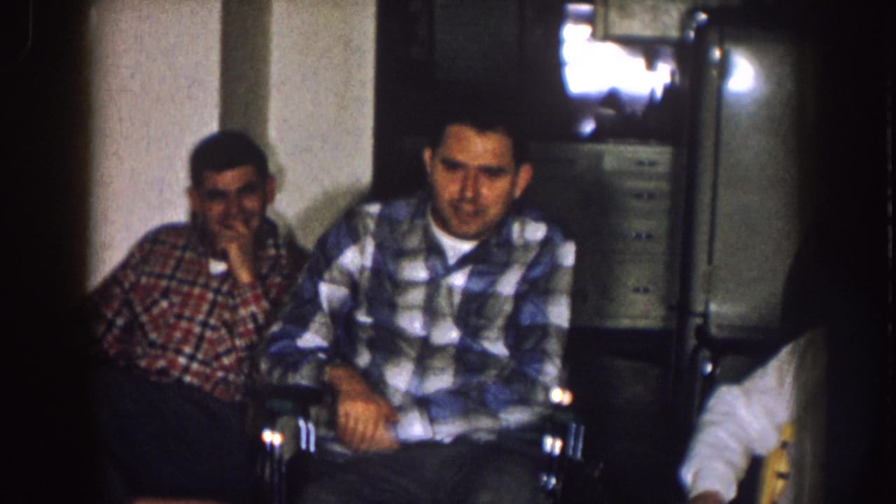 1957 Nebraska home-movie frame showing two adult men seated in a dim room, with a television and wall behind them.