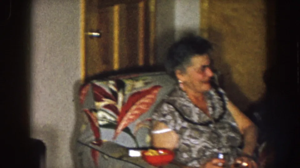 1957 Nebraska home-movie frame showing an older woman seated in profile beside a floral cushion and wood door in a dim private room.