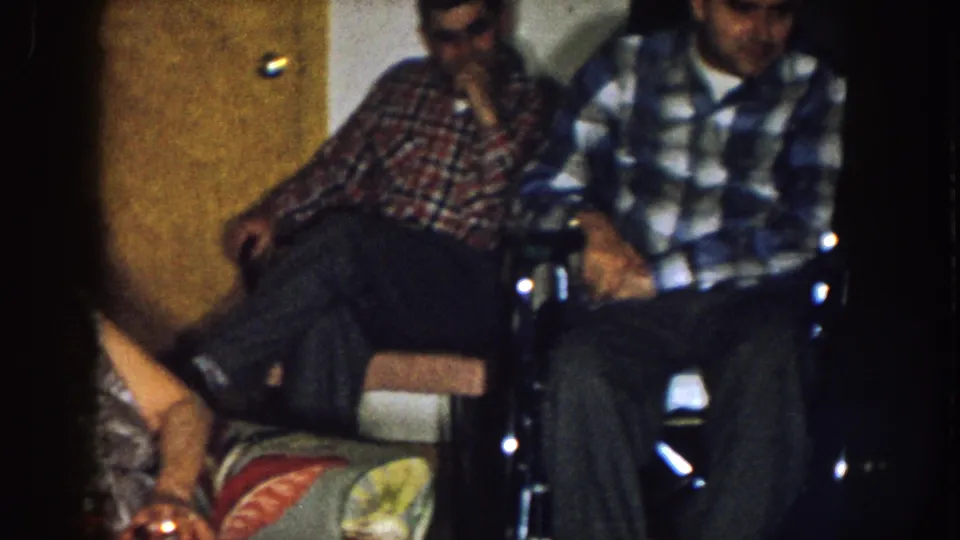 1957 Nebraska home-movie frame showing two adult men in plaid shirts seated close together in a private room with a wooden door behind them.