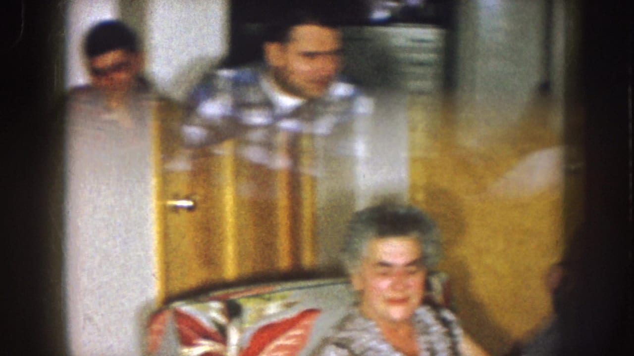 1957 Nebraska home-movie frame showing adults seated indoors near a brown door, including an older woman in the foreground.