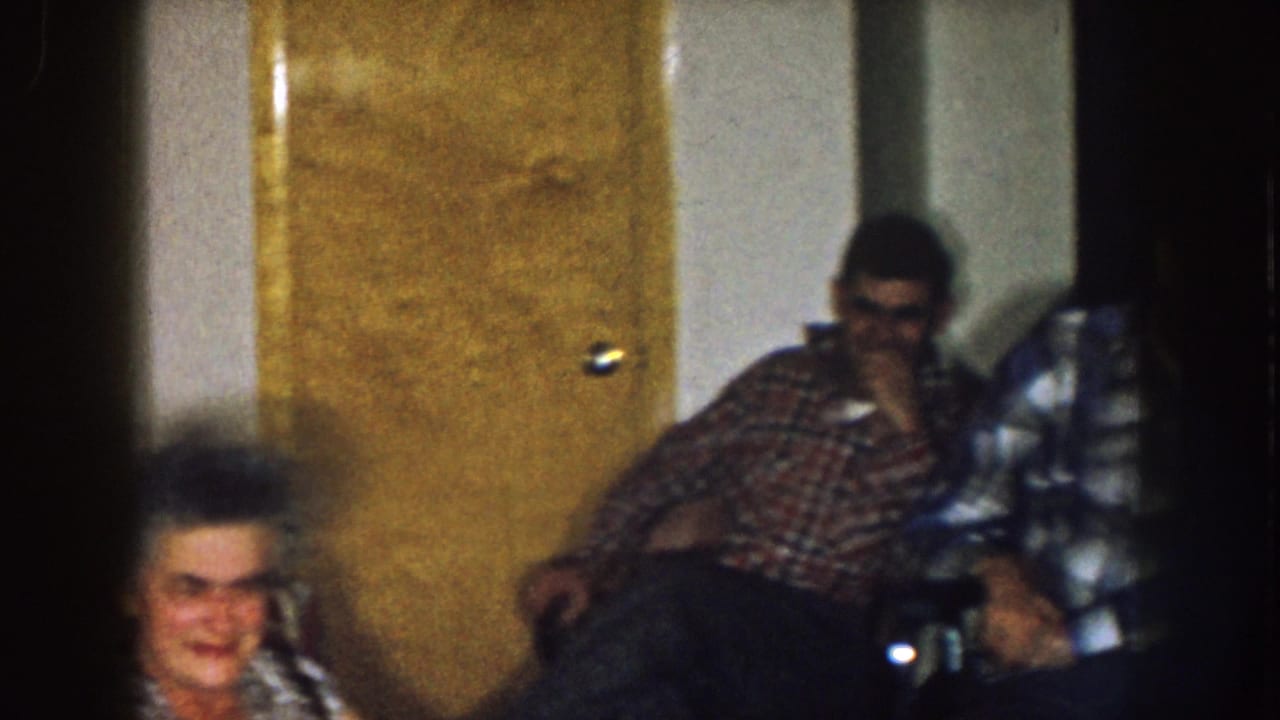 Two men sit near a brown door, with part of an older woman visible at the bottom of the frame.