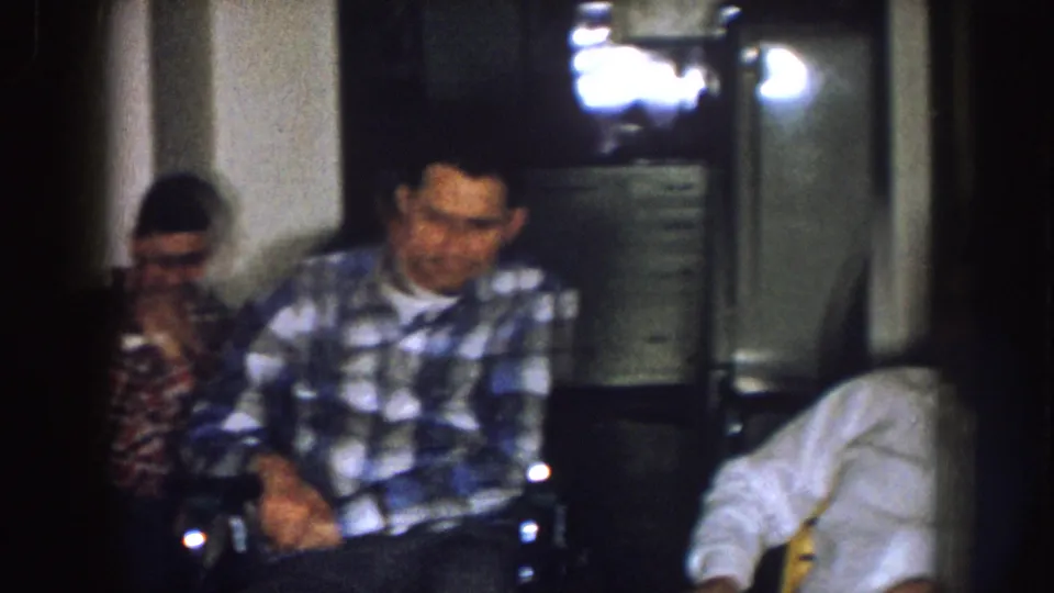 1957 Nebraska home-movie frame of two seated men indoors, one wearing a blue plaid shirt and one in a red checkered shirt.