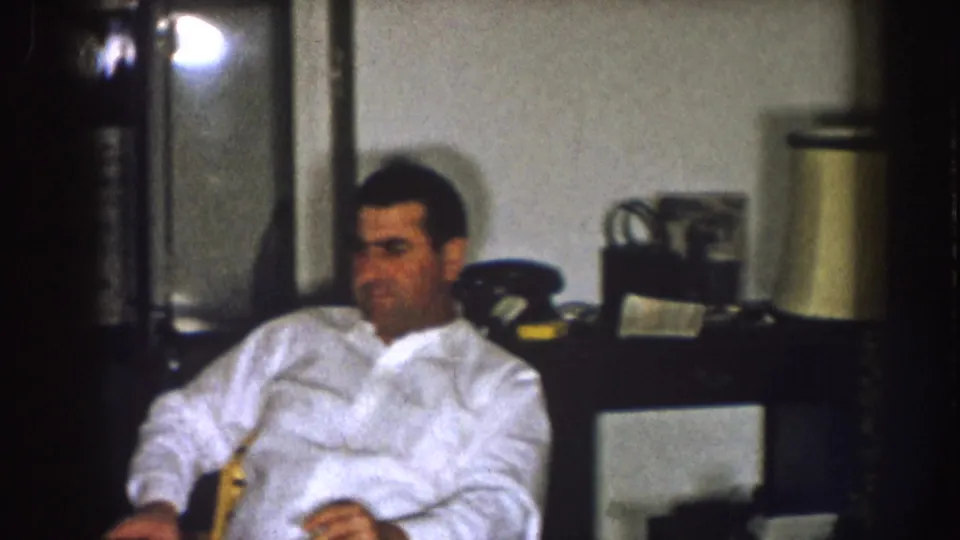 1957 Nebraska interior frame showing a seated man in a white shirt near household objects in a dim private room.