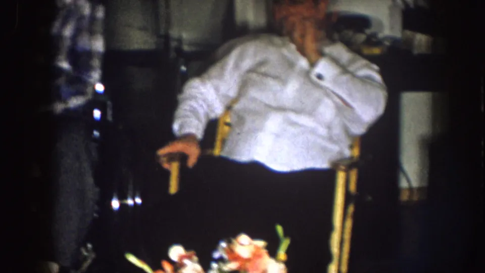 A man in a white shirt sits in a wooden chair in the living room, with dark furniture and soft 8mm exposure around him.