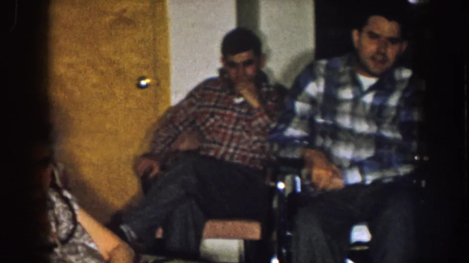 Two men sit near a wooden door in the Nebraska home interior, one in plaid clothing and the other in a checkered shirt.