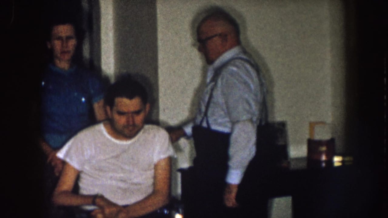 1957 Nebraska home-movie frame showing a man seated in a wheelchair while a woman and older man stand behind him in a dim interior.