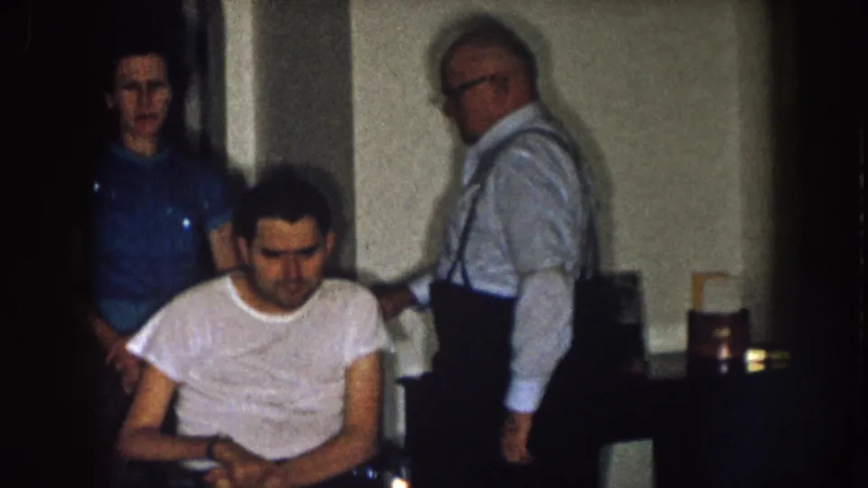 1957 Nebraska home-movie frame showing a man seated in a wheelchair while a woman and older man stand behind him in a dim interior.