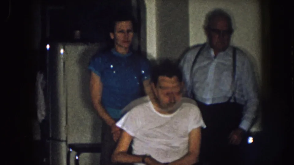 1957 Nebraska home interior with a seated man in a wheelchair, a woman in blue, an older man with suspenders, and a refrigerator behind them.