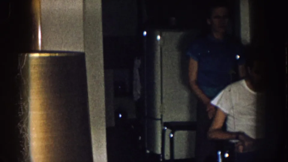 Dark 8mm frame from a Nebraska home interior, with the seated man partly obscured and two standing adults near a refrigerator.
