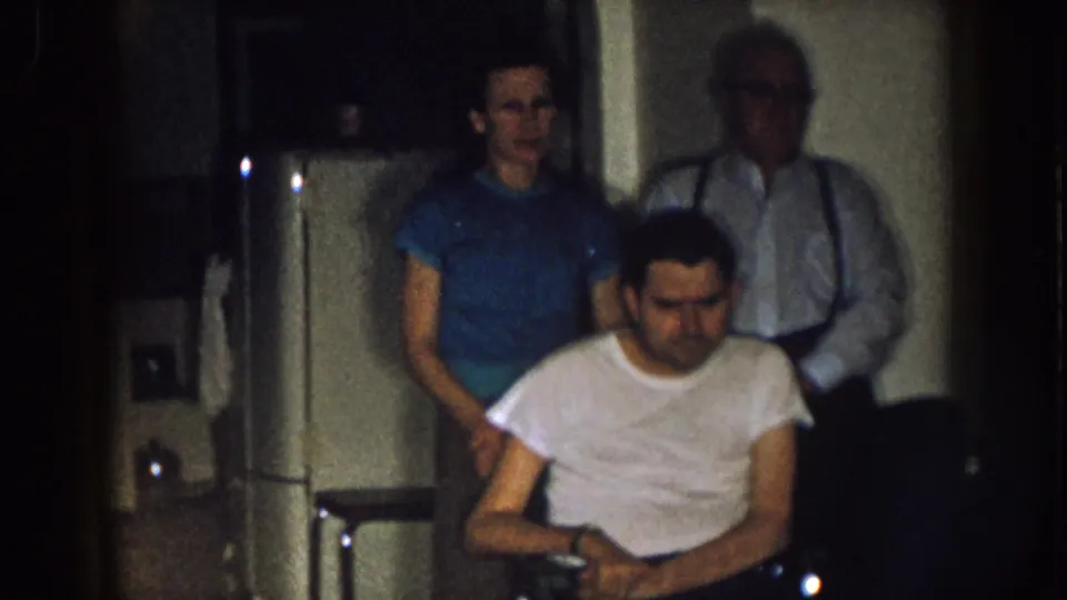 1957 Nebraska home-movie frame showing a man in a wheelchair before a woman in blue and an older man with suspenders indoors.
