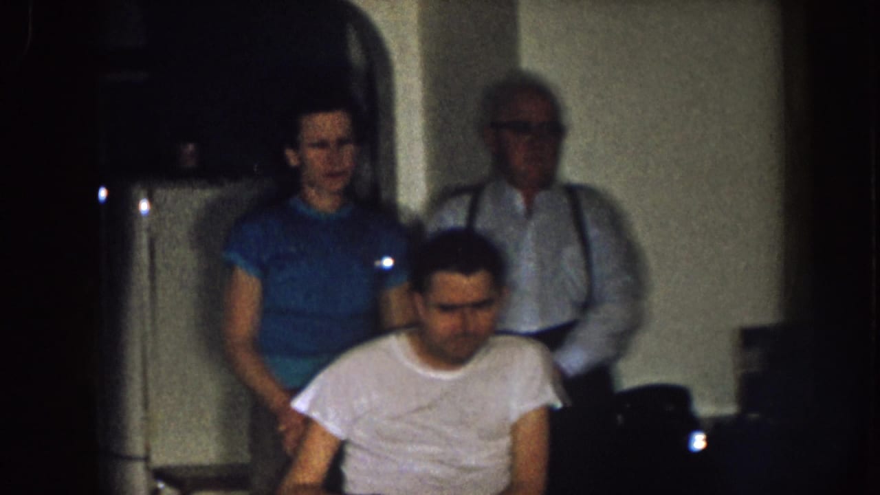 In a dim 1957 Nebraska home interior, a man sits in a wheelchair while a woman in blue and an older man in suspenders stand behind him.