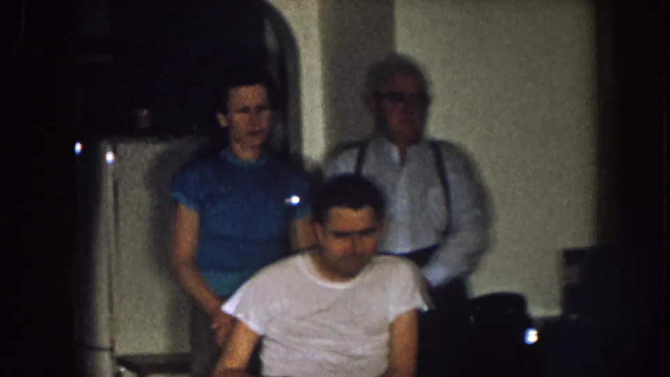 In a dim 1957 Nebraska home interior, a man sits in a wheelchair while a woman in blue and an older man in suspenders stand behind him.