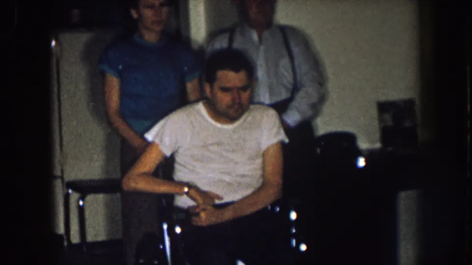 The seated man in a wheelchair looks downward as a woman and older man stand behind him in a dim Nebraska home interior.