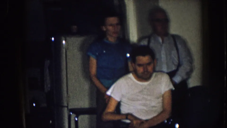 A grainy 8mm frame shows the wheelchair user seated in the foreground, with two adults behind him inside a modest 1957 Nebraska home.