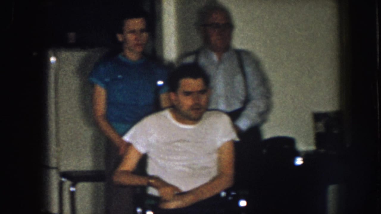 Three adult men indoors near a white refrigerator, with one seated in a white T-shirt.