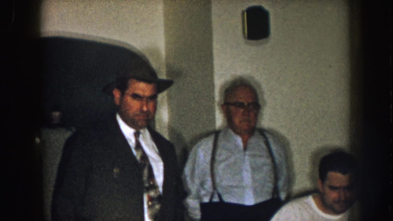 Man in dark coat and hat stands near an older man in suspenders and a seated man.