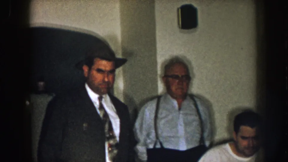1957 Nebraska indoor frame with a man in a dark coat and fedora standing beside an older man in suspenders near a seated man.