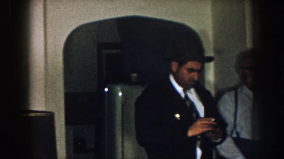 1957 Nebraska home-movie frame of a man in a dark coat and hat looking down at a small object, with an older man behind him.