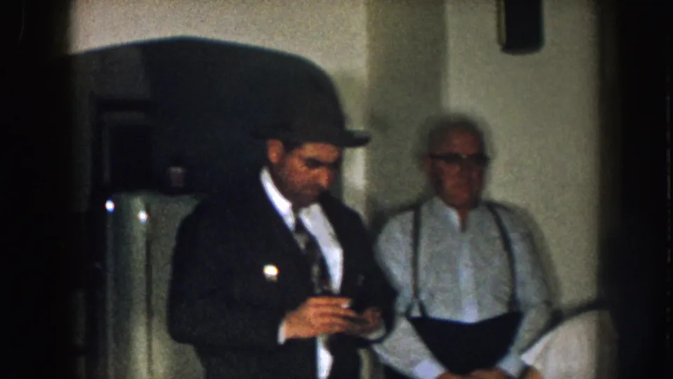 1957 Nebraska frame showing a man in a dark coat and fedora handling a small object, with an older man in suspenders behind him.