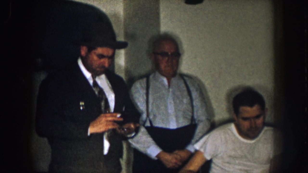 Three adult men in a dim room, with one seated in a wheelchair and two standing nearby.
