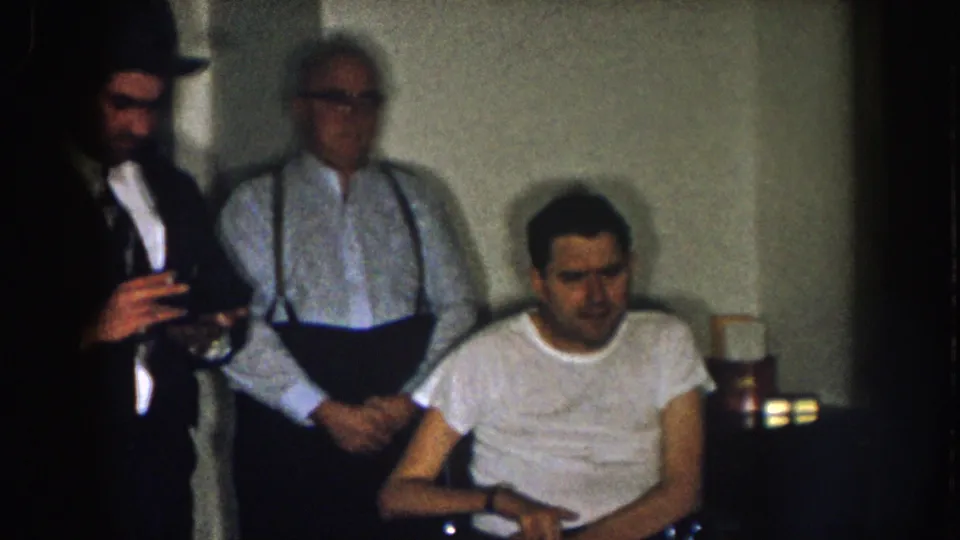 1957 Nebraska frame with a suited man holding a glass, an older man in suspenders, and a seated man in a wheelchair.