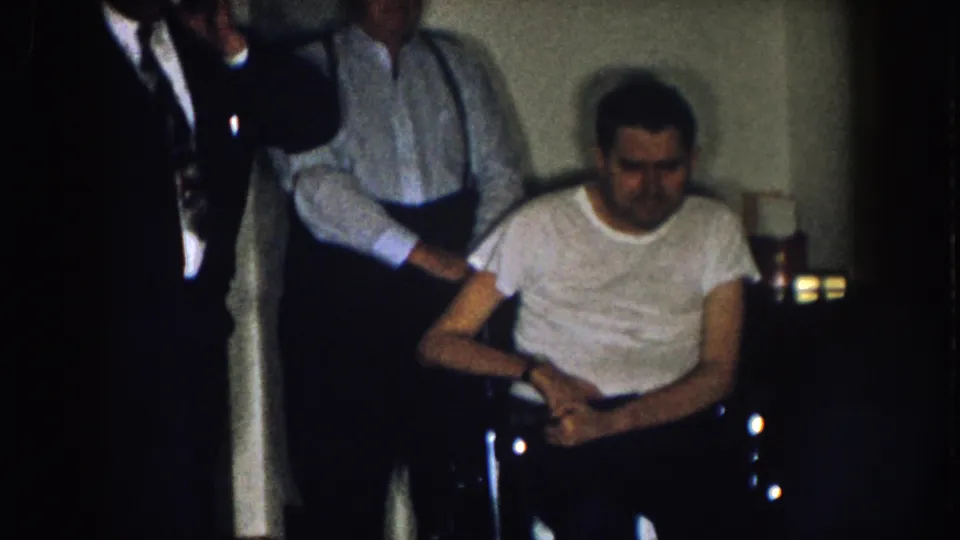 1957 Nebraska home-movie frame centering a man in a wheelchair with two adult men standing nearby in a dark room.
