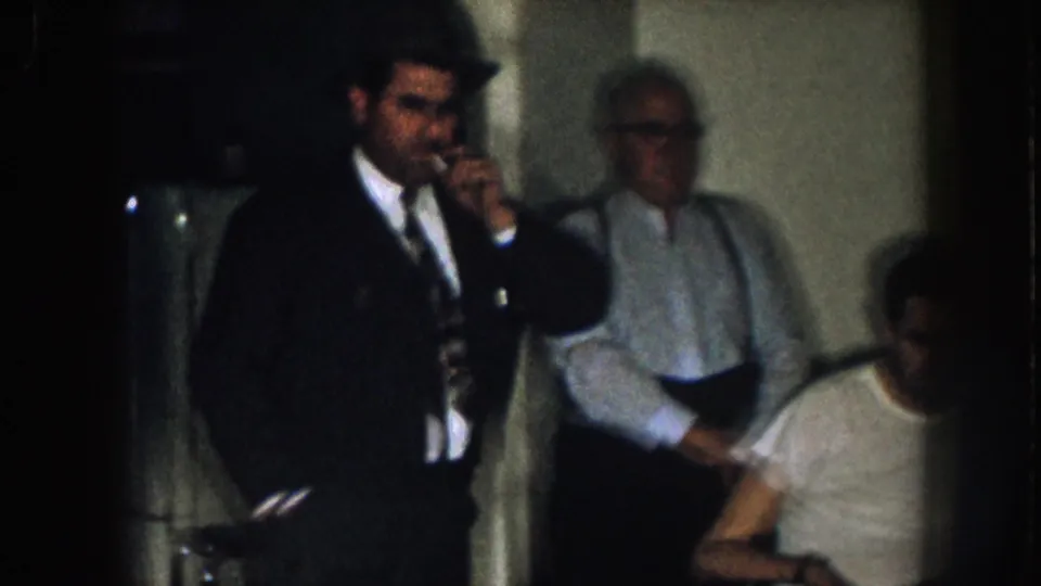 1957 Nebraska frame showing a suited man lifting a glass beside an older man, with the seated wheelchair user partly visible.