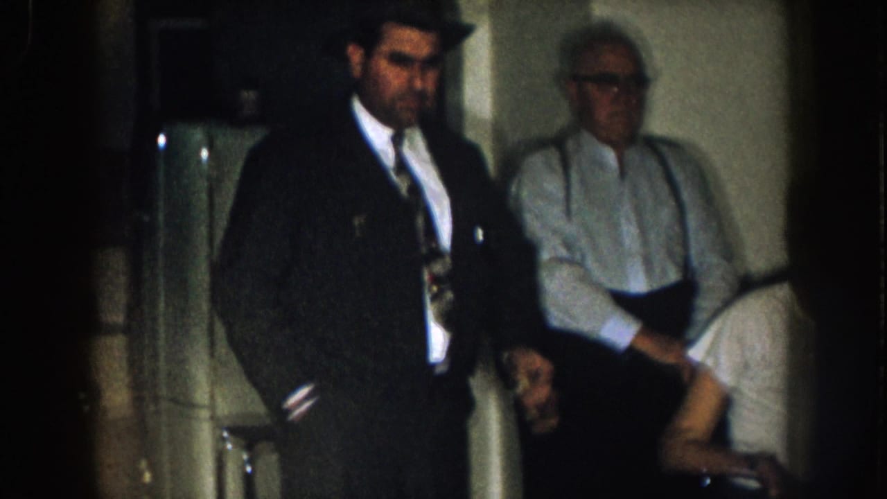 Nebraska 1957 home-movie frame showing a suited man in a fedora, an older man in suspenders, and a seated man in a dim private room.