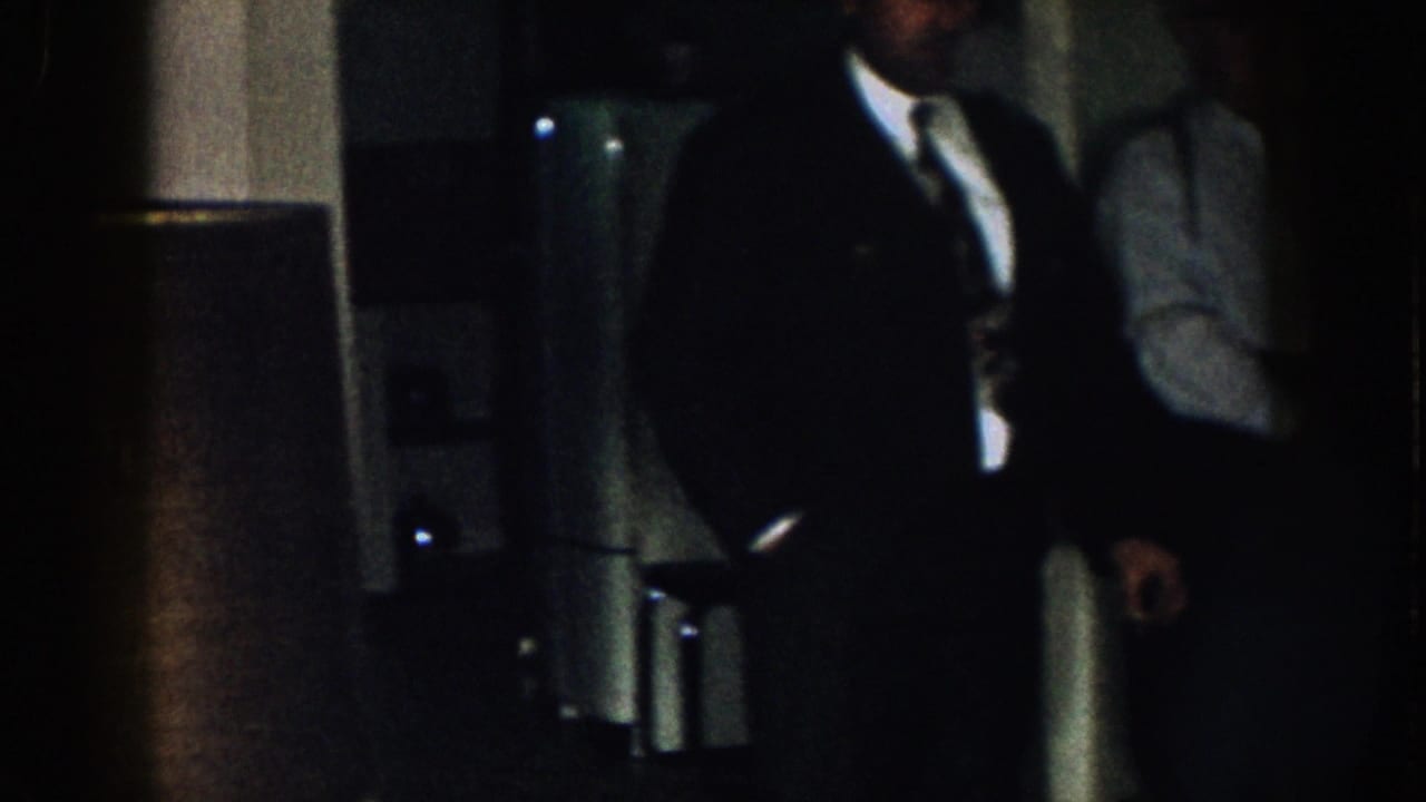 A suited man is partly cropped beside a dark doorway or cabinet in a low-lit room.
