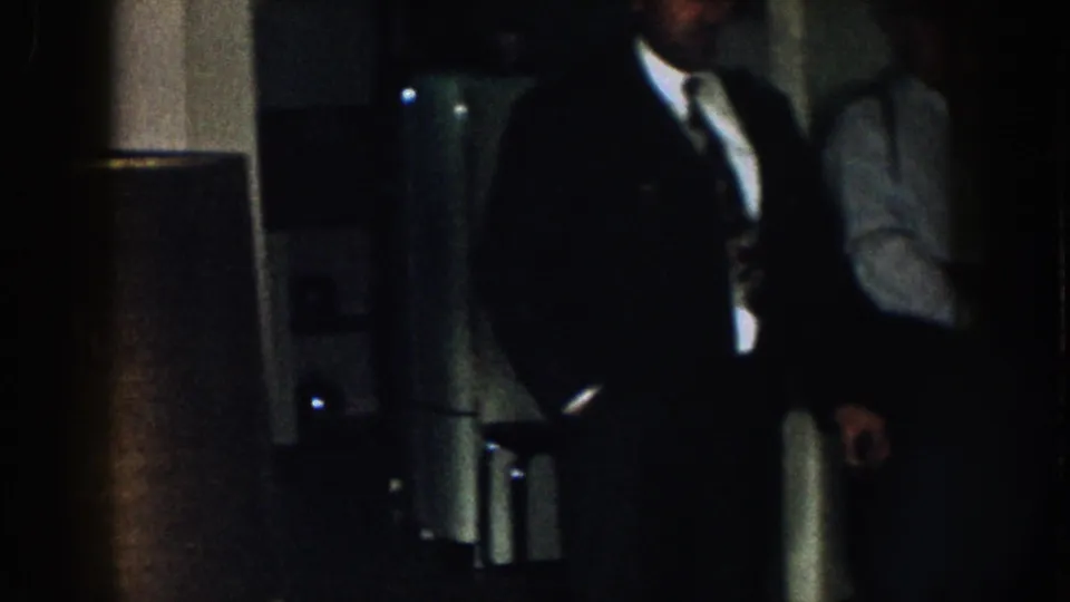 Nebraska 1957 frame with a dark-suited man partly cropped at right, standing near a shadowed interior fixture or doorway.