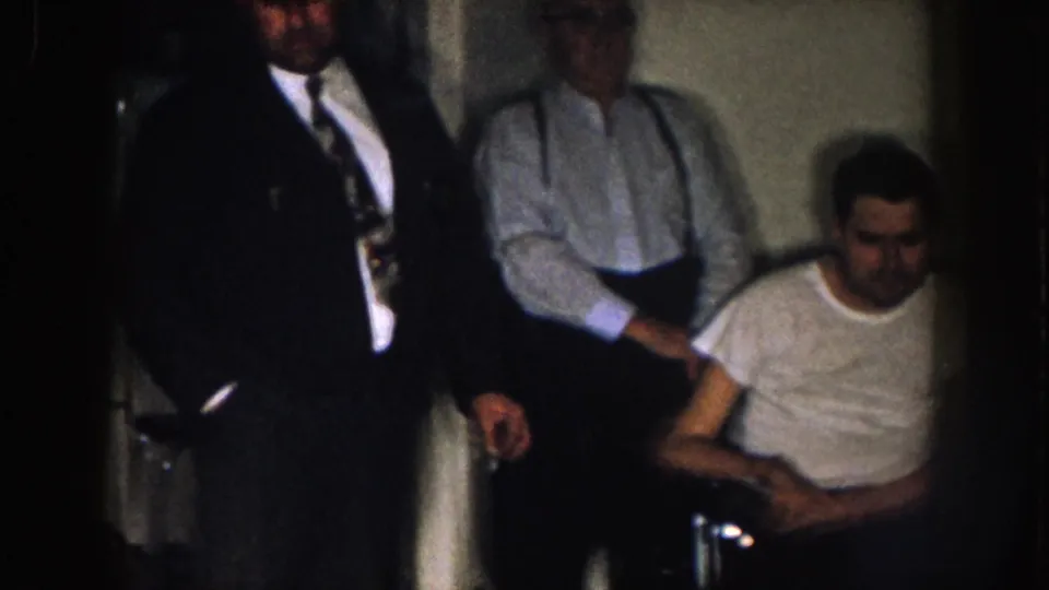 Nebraska 1957 home-movie frame showing standing adult men near a seated man in a white t-shirt, filmed in a tight private interior.