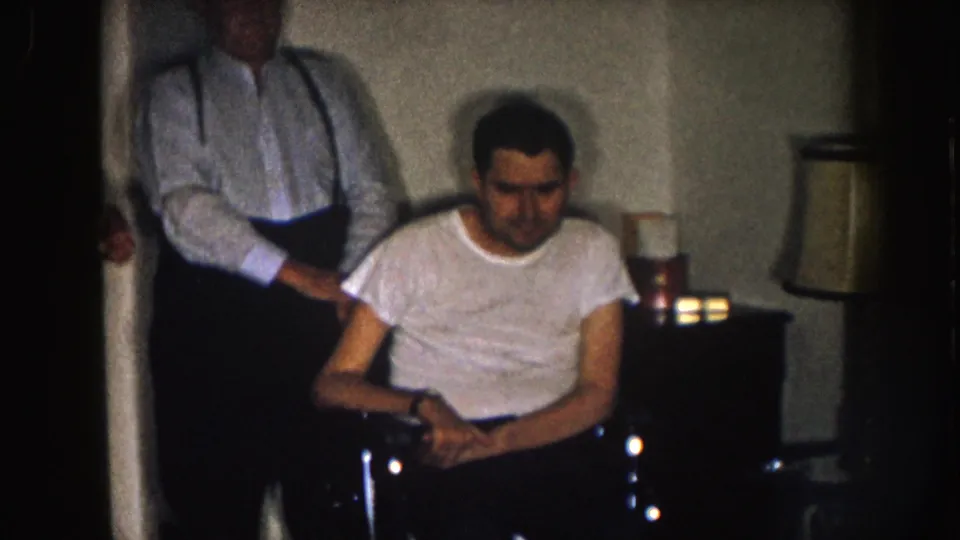 Nebraska 1957 frame showing a seated man in a white t-shirt near a table lamp, with an older man in suspenders standing behind him.