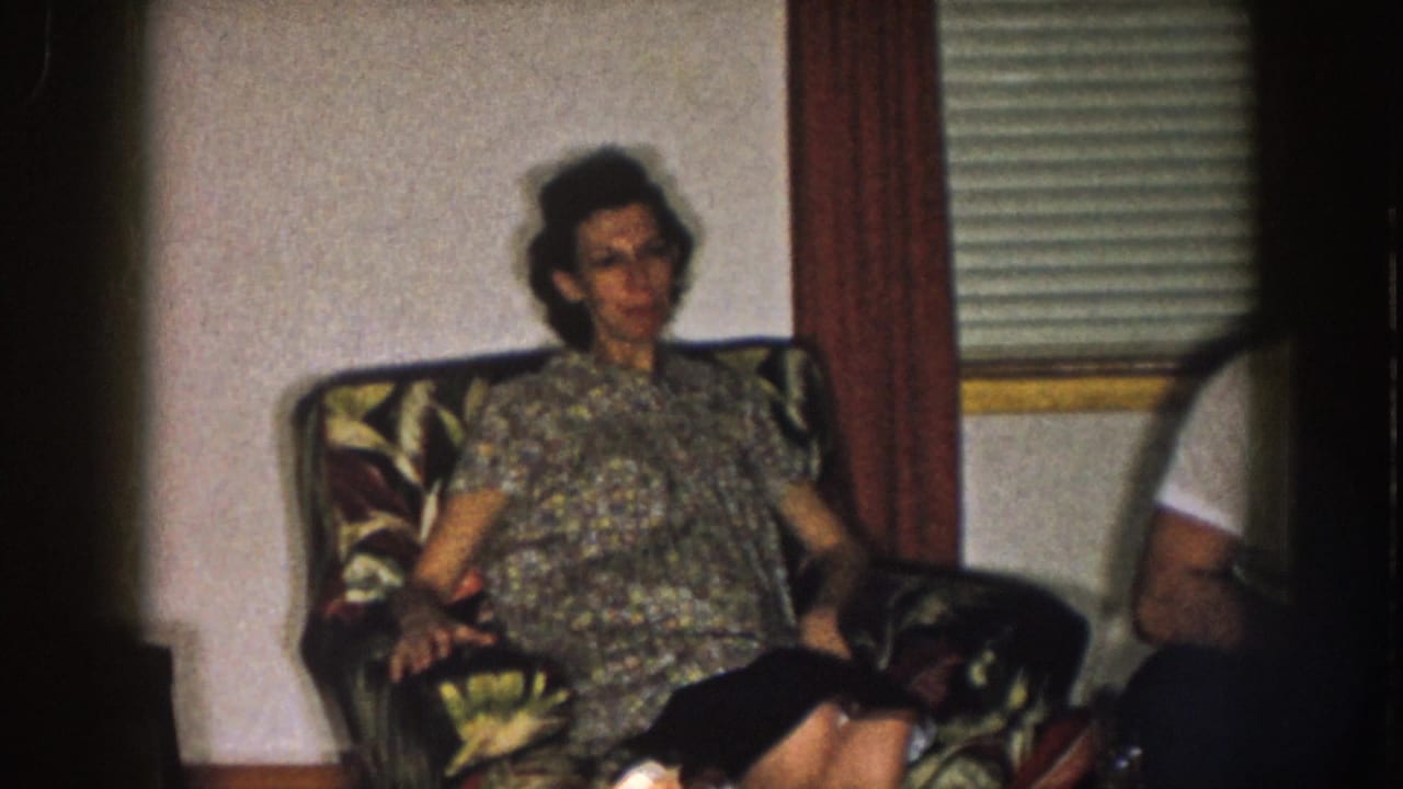 1957 Nebraska home-movie frame showing a woman in a patterned dress seated in a floral armchair.