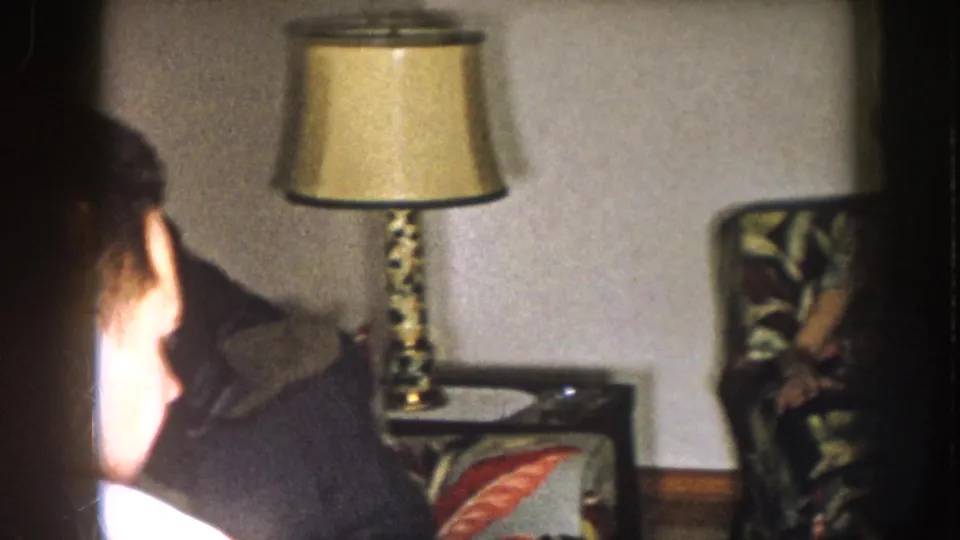 Handheld 1957 Nebraska frame with adults seated near a table lamp and floral chairs in a private living room.