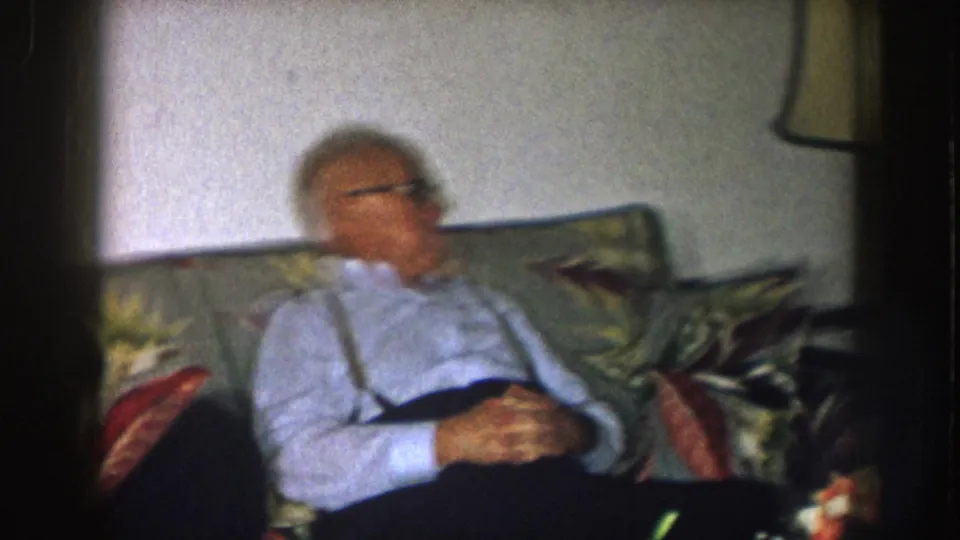 1957 Nebraska home-movie frame showing an older man in suspenders seated on a floral couch in a living room.