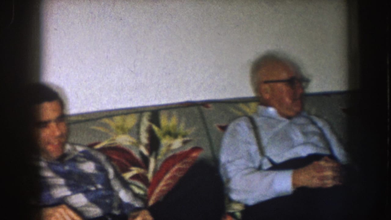 1957 Nebraska home-movie frame showing two adult men seated on a floral couch in a living room interior.