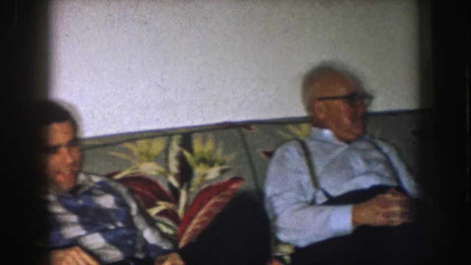 1957 Nebraska home-movie frame showing two adult men seated on a floral couch in a living room interior.