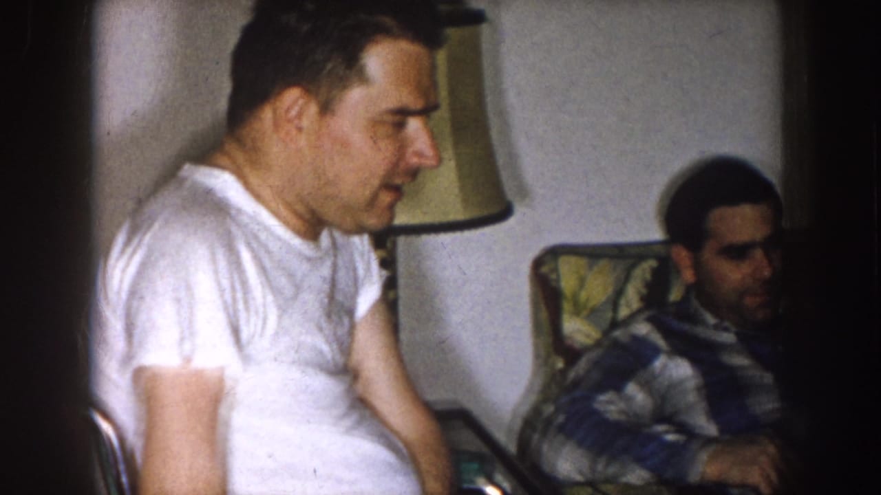 Man in a white t-shirt sits in the foreground with another man on a chair or couch nearby.