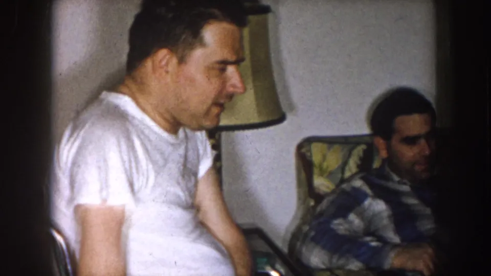 1957 Nebraska living room frame with a man in a white t-shirt in profile and another seated man at right.