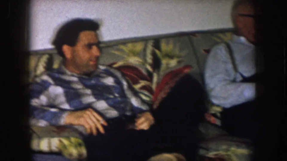 1957 Nebraska home interior with a plaid-shirted man on a floral couch and an older man partly in frame.