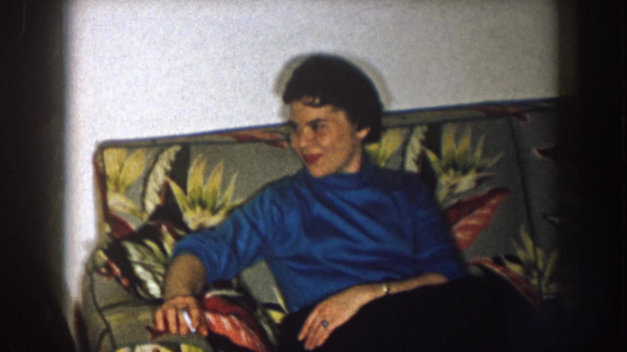 In a 1957 Nebraska home-movie frame, a woman in a blue turtleneck sits on a floral sofa and looks off camera.