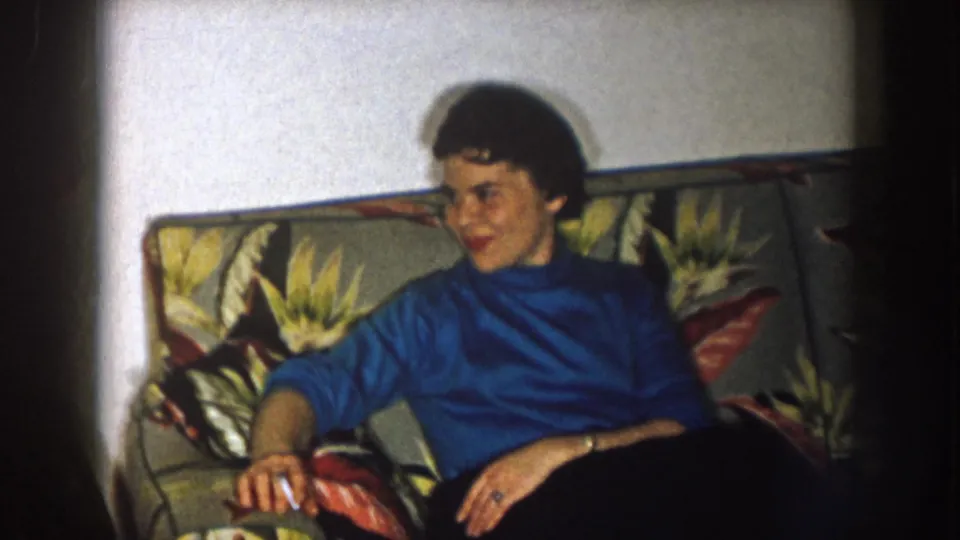 In a 1957 Nebraska home-movie frame, a woman in a blue turtleneck sits on a floral sofa and looks off camera.