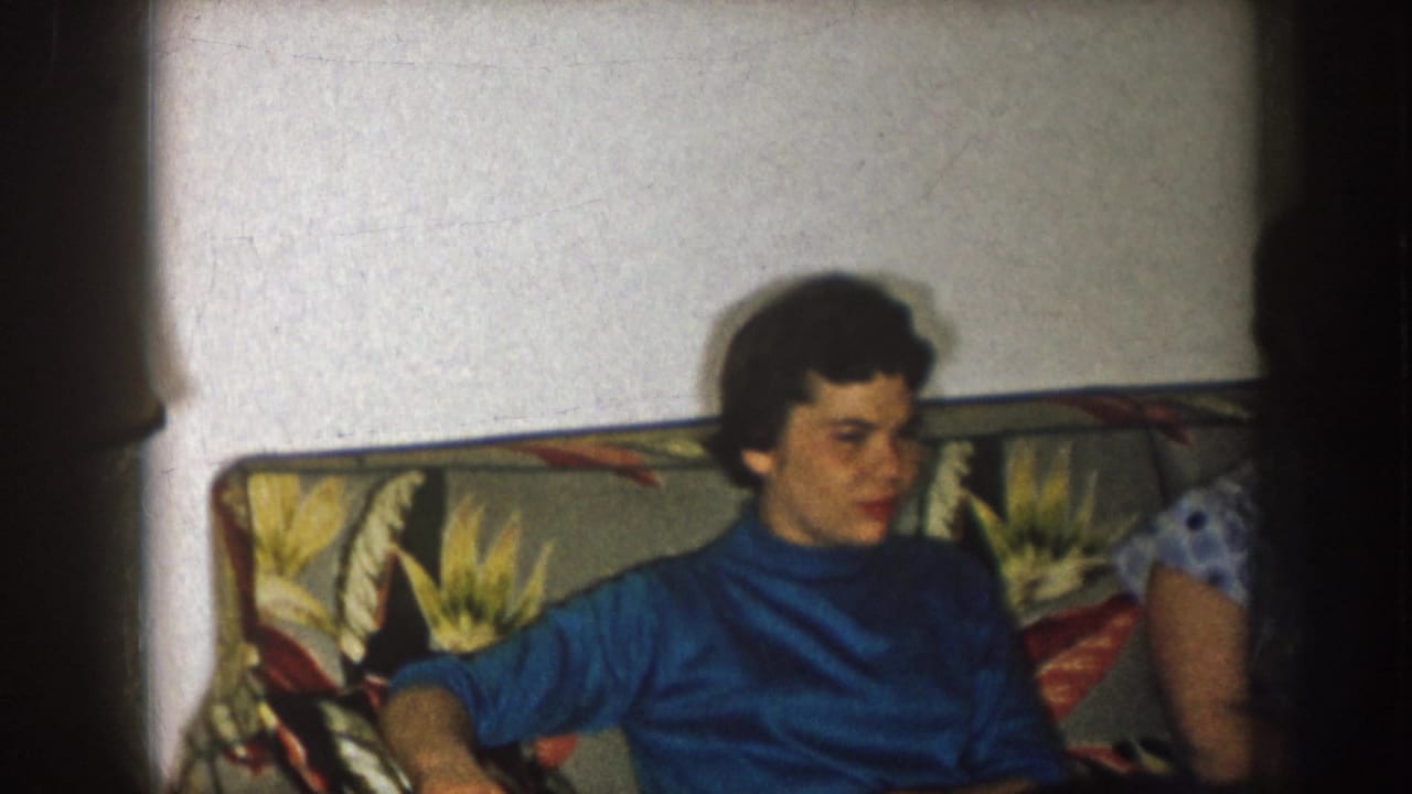 Woman in a blue turtleneck seated on a floral sofa, turned toward another person.