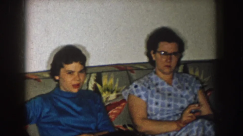Two women sit side by side on a floral sofa in a 1957 Nebraska home interior, one in a blue turtleneck and one in glasses.